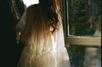 Woman in a sheer white Noémiah embroidered Tulip dress, standing by a window with sunlight streaming in.