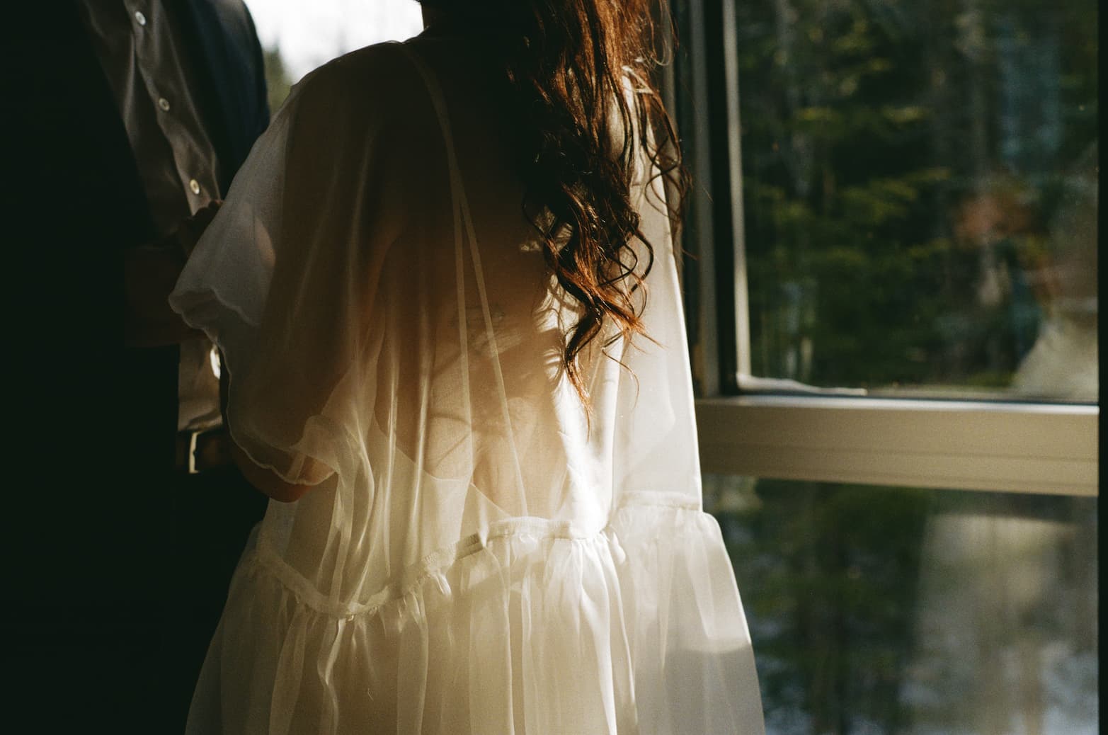 Woman in a sheer white Noémiah embroidered Tulip dress, standing by a window with sunlight streaming in.