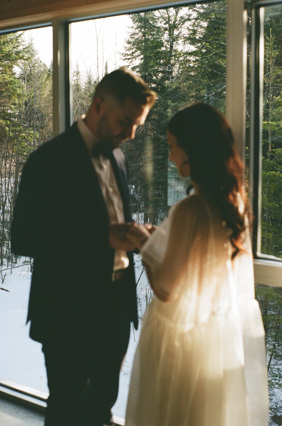 Couple standing by a large window in a forested area, with the woman wearing a Noémiah embroidered Tulip dress.