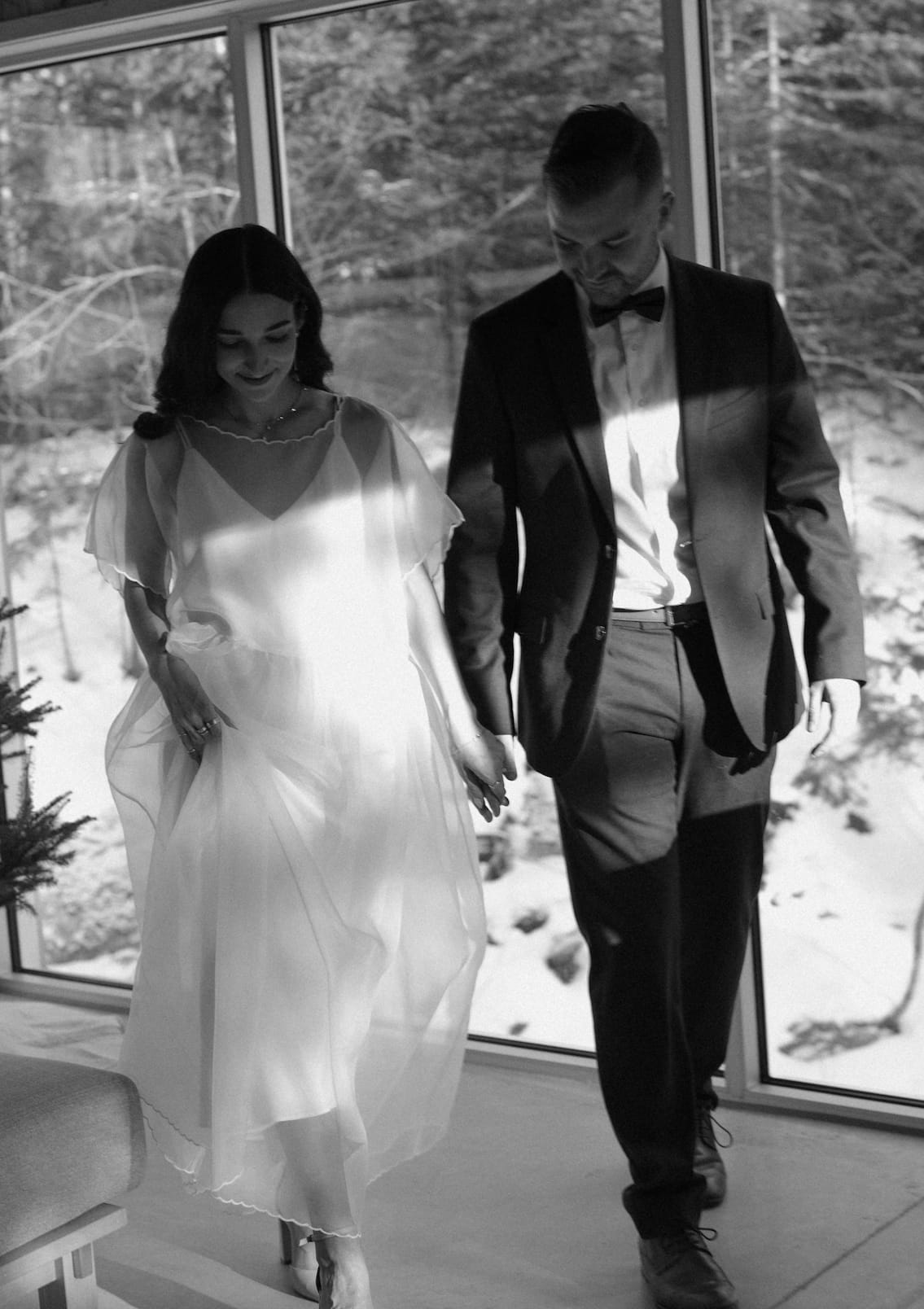 A woman in a flowing Noémiah embroidered Tulip dress walks hand-in-hand with a man in a suit by a snowy window.