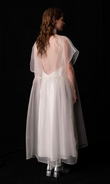 A person stands against a black background, wearing the Embroidered Tulip Dress by Noémiah, which is a flowing, sheer silk organza design paired with silver boots. The oversized dress features short, translucent sleeves and a low back. Their wavy hair falls past their shoulders.