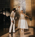 A couple dances joyfully in a sunlit hallway. The man wears a beige suit, and the woman, in a flowing Embroidered Tulip Dress by Noémiah crafted from silk organza, holds a bouquet of flowers. They twirl together on a checkered floor, surrounded by light and shadows.