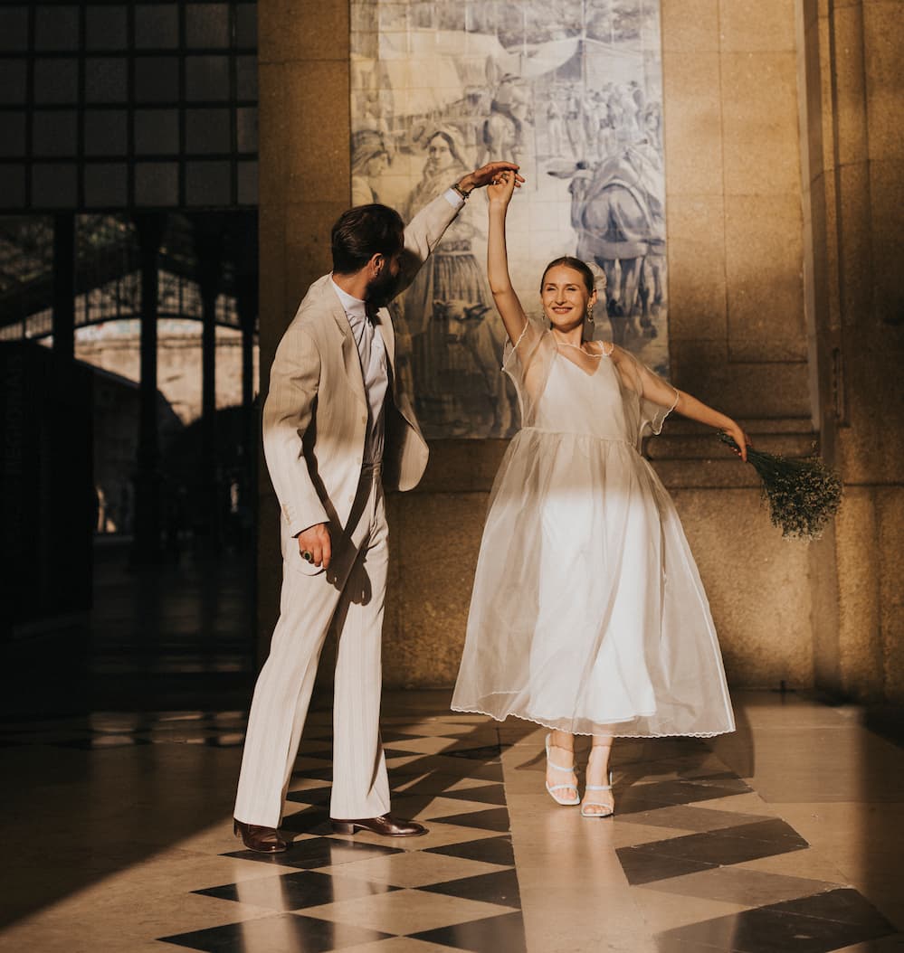 A couple dances joyfully in a sunlit hallway. The man wears a beige suit, and the woman, in a flowing Embroidered Tulip Dress by Noémiah crafted from silk organza, holds a bouquet of flowers. They twirl together on a checkered floor, surrounded by light and shadows.