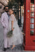 A man in a pastel suit and sunglasses stands next to a woman wearing the Embroidered Tulip Dress by Noémiah, holding a bouquet. They pose beside a red British-style phone booth on a cobblestone street, with trees and buildings in the background.