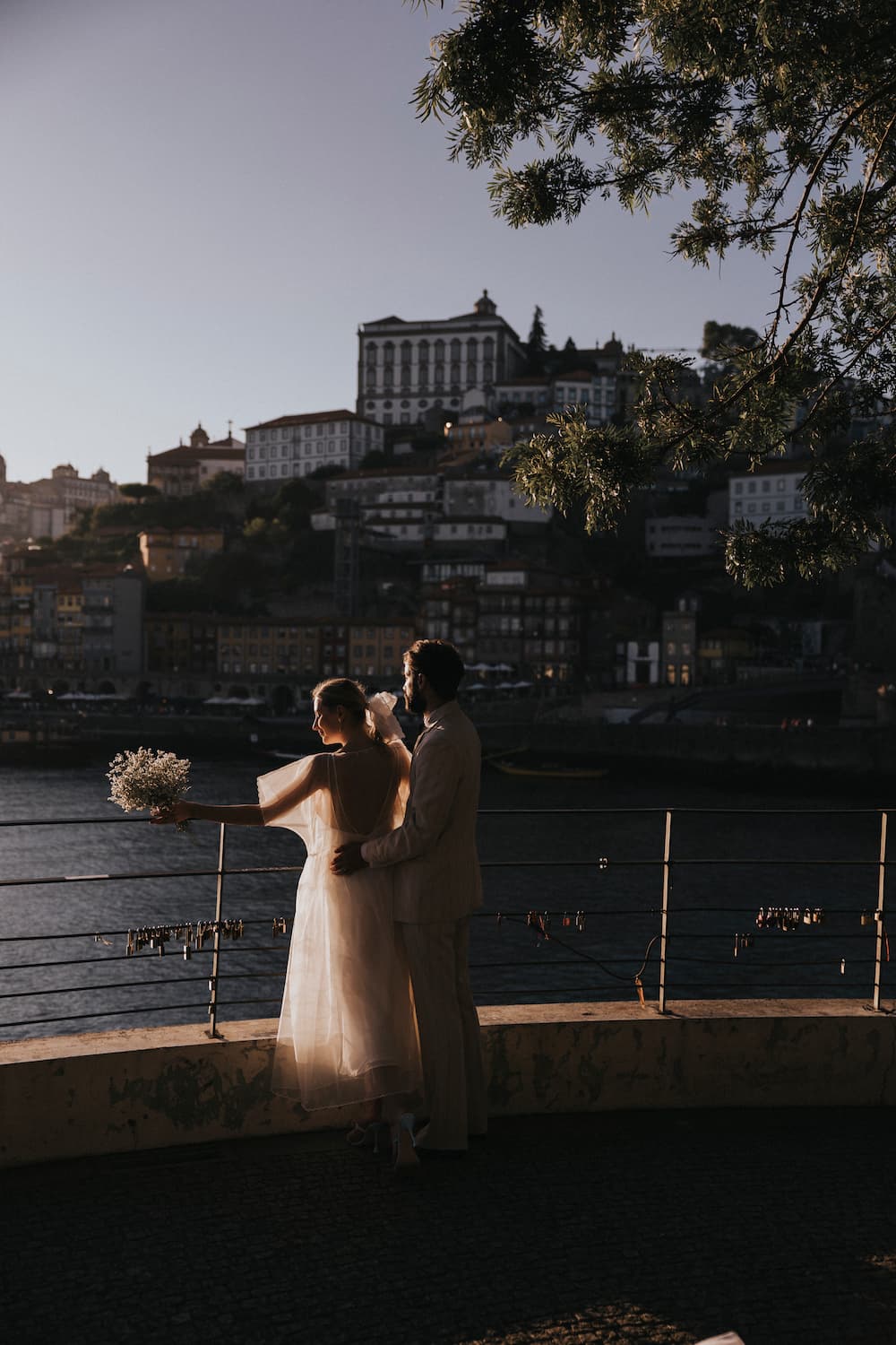 A couple in wedding attire stands by a railing overlooking a river at sunset. The bride, wearing the Noémiah Embroidered Tulip Dress, holds a bouquet. They face a scenic hillside town with historic buildings as tree branches frame the enchanting scene.