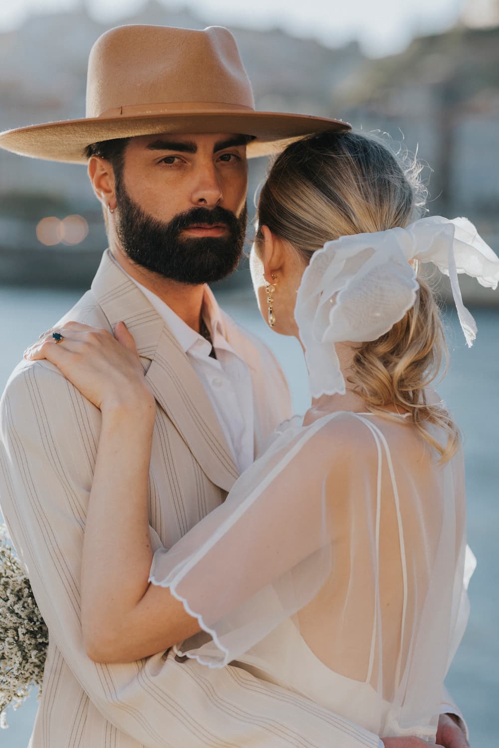 A man with a beard and a wide-brimmed hat embraces a woman. She's wearing the Embroidered Tulip Dress by Noémiah, an oversized, sheer white dress crafted in silk organza, her hair tied back with a ribbon. They stand by a body of water, with blurred buildings in the background.