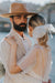 A man with a beard and a wide-brimmed hat embraces a woman. She's wearing the Embroidered Tulip Dress by Noémiah, an oversized, sheer white dress crafted in silk organza, her hair tied back with a ribbon. They stand by a body of water, with blurred buildings in the background.