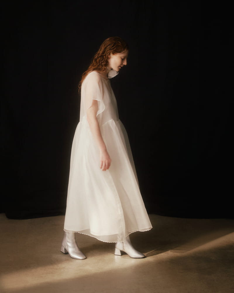 A person with long hair, elegantly clad in a flowing Embroidered Tulip Dress by Noémiah and silver boots, strides across a dimly lit room with a dark background.