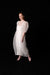 A person with light skin and red hair stands against a black background, wearing the Noémiah Céline Silk Satin Dress — Bridal Edit with puffed short sleeves and sheer white fabric, paired with pale pink heels.