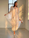 A person in the elegant Esperanza Maxi Dress — Bridal Edit by Noémiah poses gracefully in a sunlit room, with silk straps enhancing the delicate quality of the sheer white dress. One leg is slightly bent, metallic heels shimmer, and sunlight pours through the window, casting shadows on the floor and highlighting the ethereal charm of this handmade-to-order ensemble.