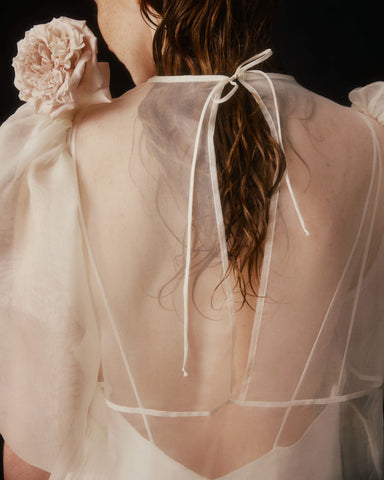 A person with wet hair is adorned in the Esperanza Maxi Dress — Bridal Edit by Noémiah, a handmade-to-order piece featuring delicate silk straps and a tied ribbon at the back, with a fabric flower on the shoulder. The image captures the back and part of their arm against a dark background.