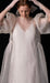 Wearing the Esperanza Maxi Dress from Noémiah's Bridal Edit, a person stands against a black background. The gown features sheer, flowing white fabric, puffed sleeves, and transparent gloves. With hands raised to their head, they look downwards in contemplative grace. The elegance of the dress highlights its handmade-to-order craftsmanship.