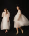 Two women are adorned in Esperanza Maxi Dresses from the Bridal Edit by Noémiah, featuring semi-transparent, ethereal white fabric with voluminous sleeves and skirts. They stand against a dark background, their delicate handmade-to-order dresses imparting an enchanting and dreamlike aura as they face different directions in the slightly blurred image.