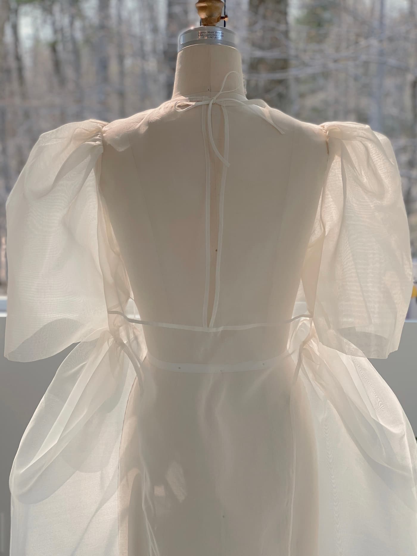 A dress form showcases the Esperanza Maxi Dress — Bridal Edit by Noémiah, featuring a sheer, ethereal design with puffed sleeves and silk straps tied at the back. Sunlight filters through trees in the background, creating a soft and delicate atmosphere. Each of these exquisite dresses is handmade to order, ensuring meticulous attention to detail and craftsmanship.