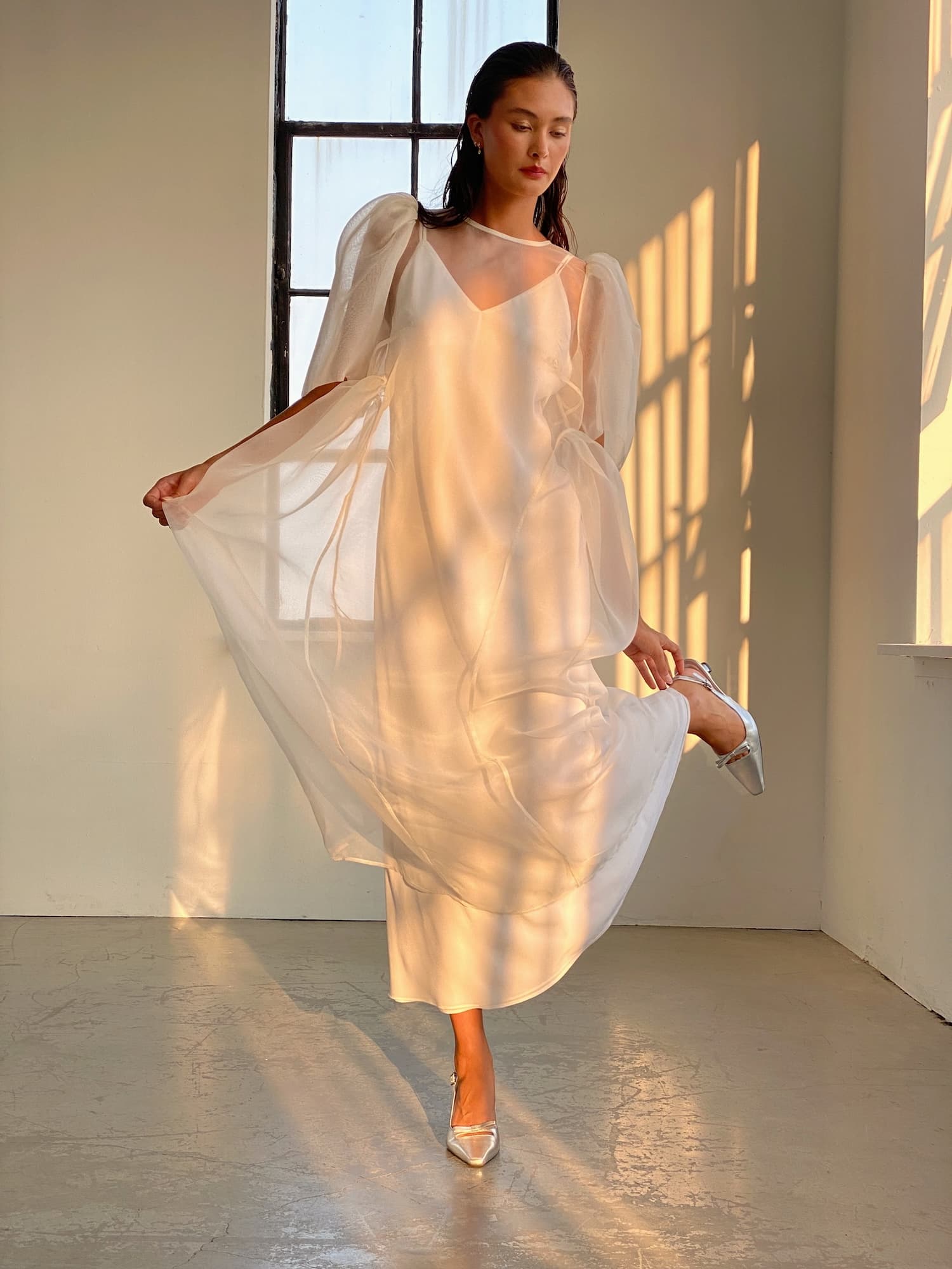 A person in the elegant Esperanza Maxi Dress — Bridal Edit by Noémiah poses gracefully in a sunlit room, with silk straps enhancing the delicate quality of the sheer white dress. One leg is slightly bent, metallic heels shimmer, and sunlight pours through the window, casting shadows on the floor and highlighting the ethereal charm of this handmade-to-order ensemble.