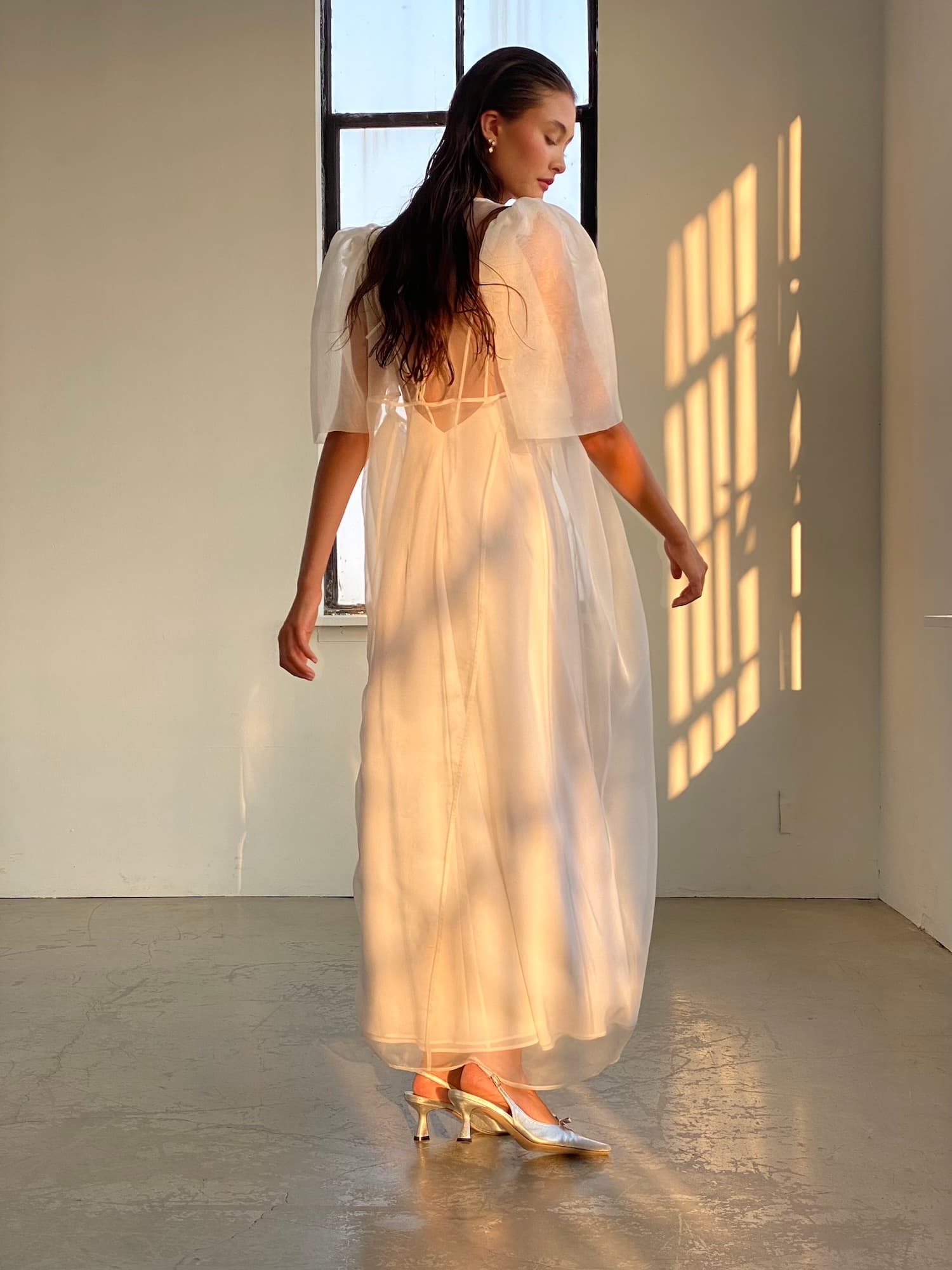 A woman wearing the elegant Esperanza Maxi Dress — Bridal Edit by Noémiah stands in a softly lit room, facing sideways. Sunlight filters through a large window, casting gentle shadows on the floor and wall. Her hair cascades loosely down her back, and she completes the look with graceful, pointed high heels.