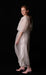 A woman stands against a black background, wearing the Esperanza Maxi Dress — Bridal Edit by Noémiah, featuring delicate silk straps over a solid white outfit. Handmade to order, she gazes at the camera with a slight smile. Her hair is slicked back, and she completes her look with pink heels.