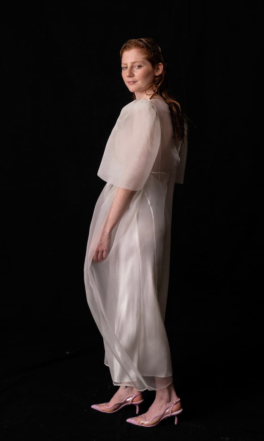 A woman stands against a black background, wearing the Esperanza Maxi Dress — Bridal Edit by Noémiah, featuring delicate silk straps over a solid white outfit. Handmade to order, she gazes at the camera with a slight smile. Her hair is slicked back, and she completes her look with pink heels.