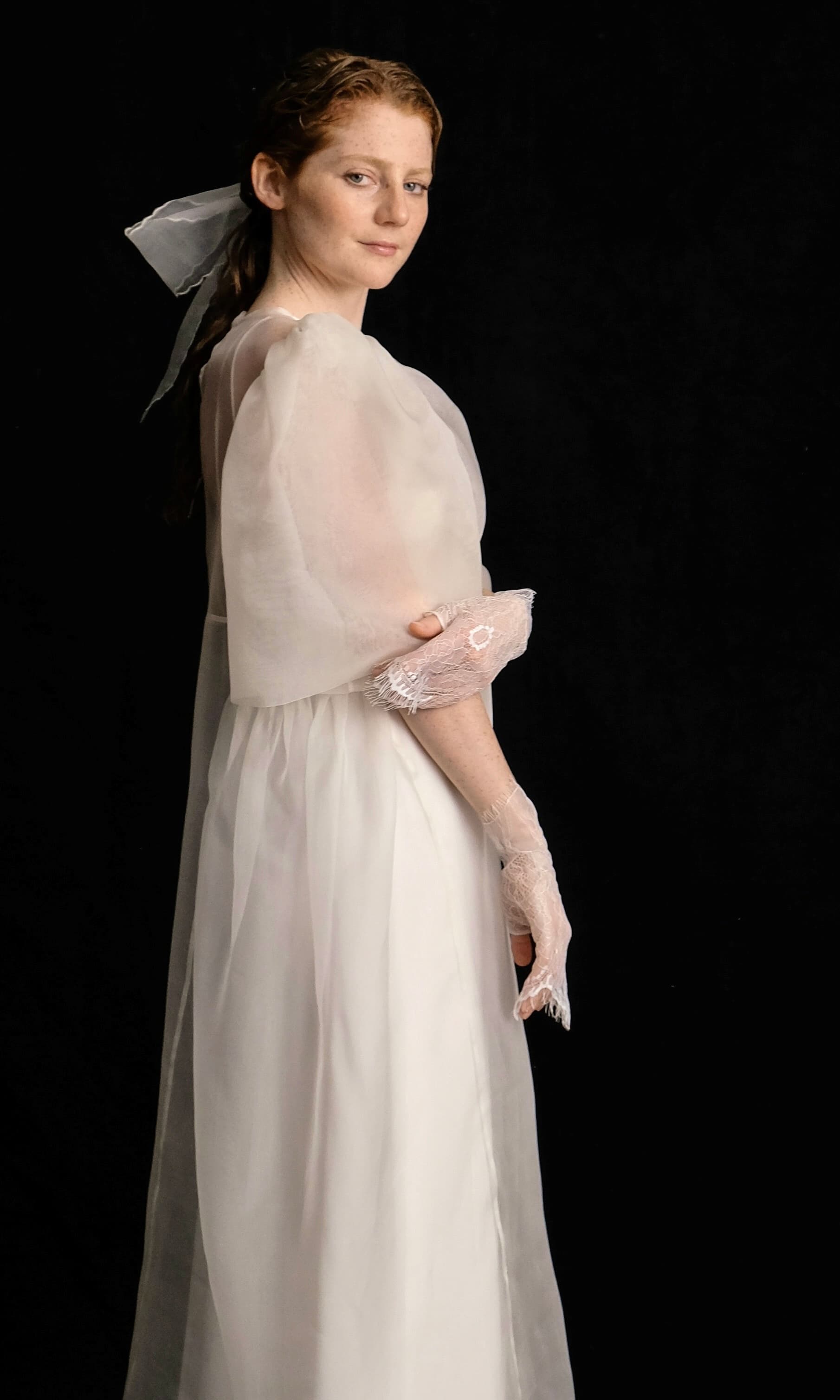 A woman wearing the Esperanza Maxi Dress from Noémiah's Bridal Edit stands against a black background. The vintage-style gown features delicate silk straps, complemented by lace gloves and a sheer fabric bow in her hair. Her expression is calm as she gazes towards the camera.