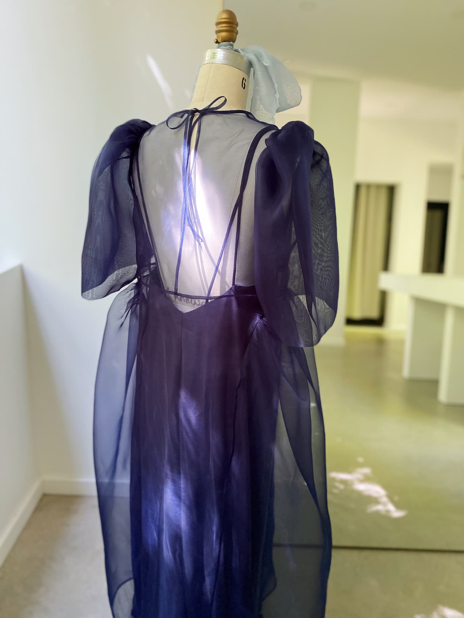 A dress form showcases the Esperanza Maxi Dress — Classic by Noémiah, a sheer, deep blue gown crafted from silk organza with puffed sleeves and an elegant cutout. Positioned in a brightly lit room, sunlight filters through to highlight the fabric's texture. The room exudes a minimalist, contemporary style.