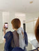 Someone with long, auburn hair takes a mirror selfie. They are dressed in the Classic Esperanza Maxi Dress by Noémiah and have styled their hair with a large, translucent white bow. The room is brightly lit and features minimalist decor.