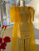 The Esperanza Maxi Dress — Classic by Noémiah, crafted from sheer silk organza in a bright yellow hue and featuring puffed sleeves, is elegantly displayed on a dress form. Sunlight filtering through a window casts soft shadows, while red flowers in the foreground accentuate the garment's elegant cutout design.