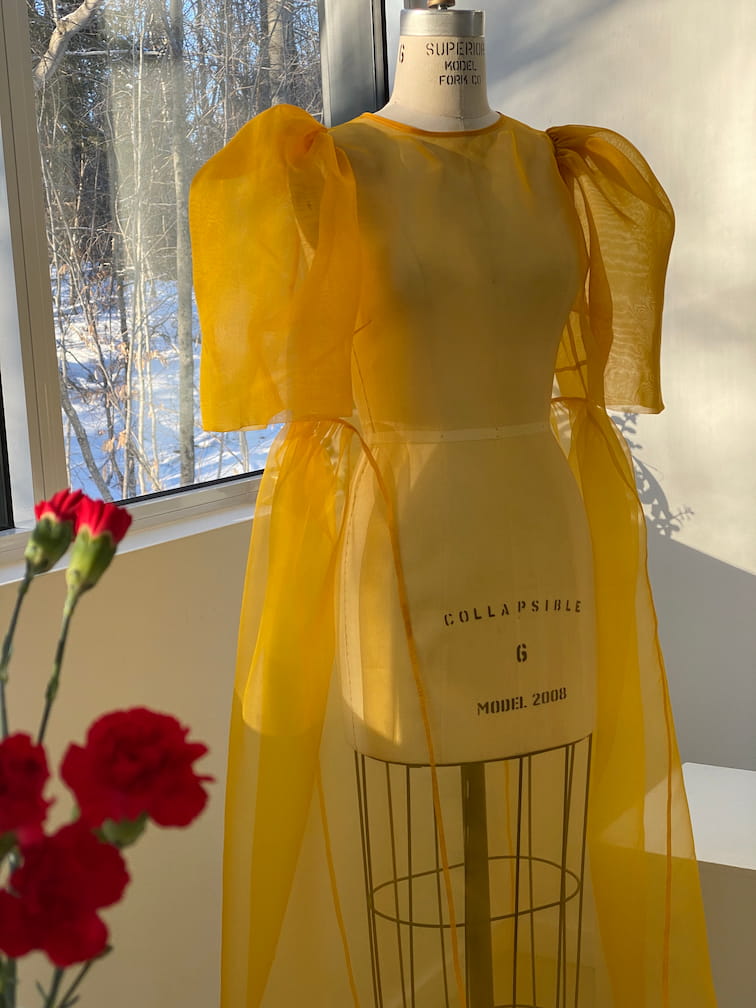 The Esperanza Maxi Dress — Classic by Noémiah, crafted from sheer silk organza in a bright yellow hue and featuring puffed sleeves, is elegantly displayed on a dress form. Sunlight filtering through a window casts soft shadows, while red flowers in the foreground accentuate the garment's elegant cutout design.