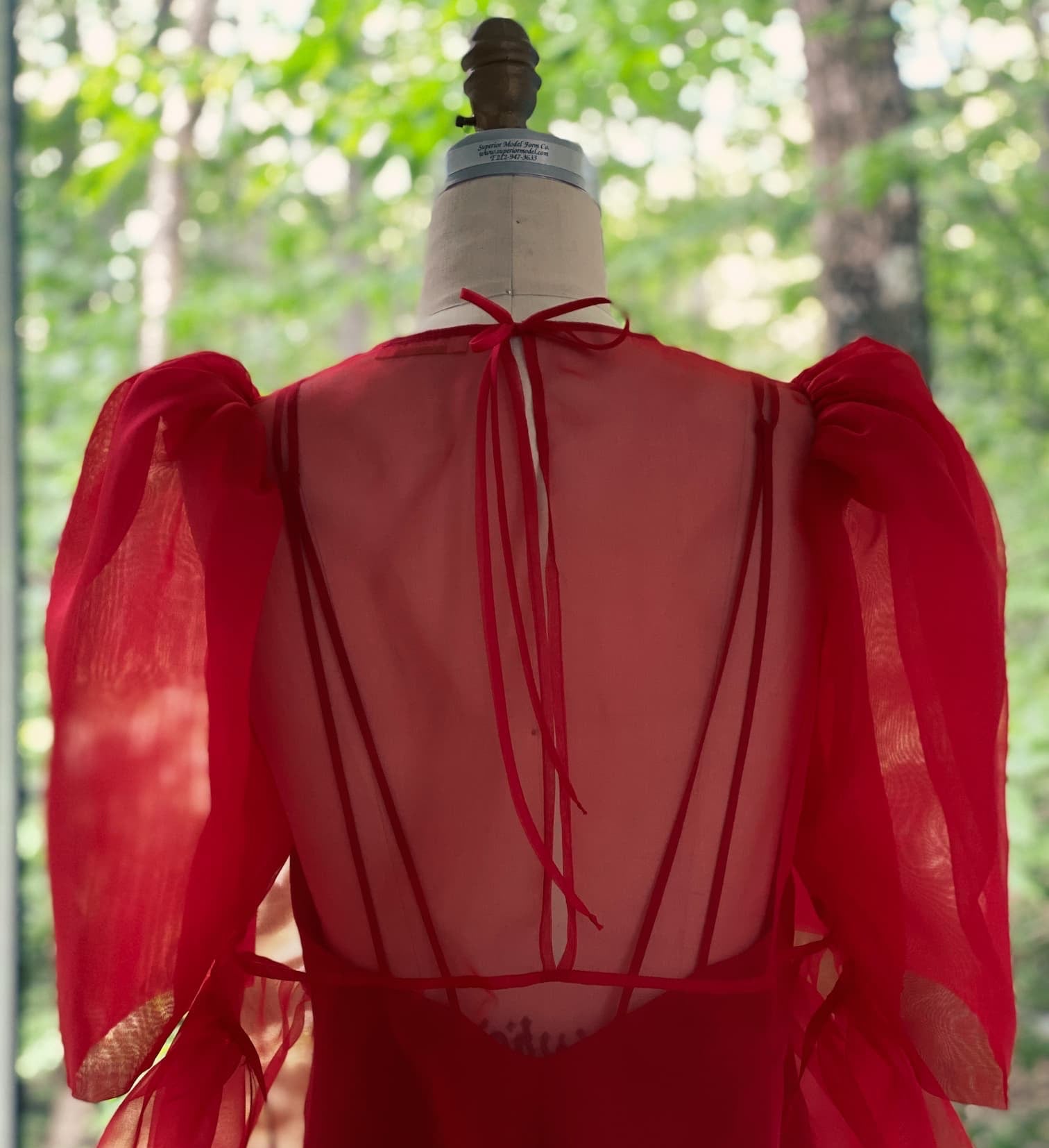 The Esperanza Midi Dress — Organza Silk by Noémiah, featuring sheer organza, puffed sleeves, and a low open back, is displayed on a mannequin in front of a window with lush green trees outside.