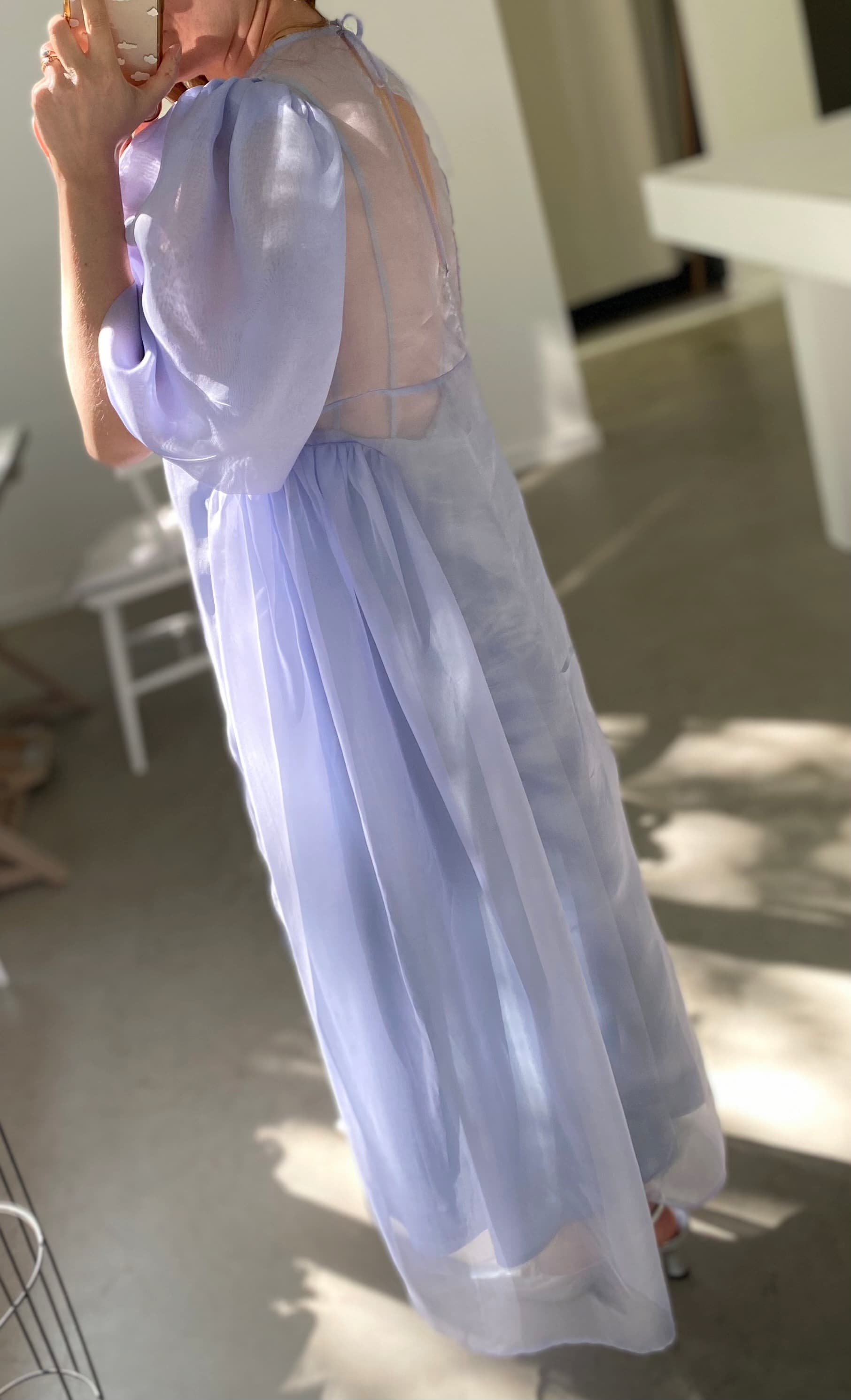 A person in the elegant Esperanza Maxi Dress — Classic by Noémiah, featuring puffed sleeves and a sheer lavender design, stands indoors near a sunlit area. They are holding a glass to their face with their back partially turned towards the camera, while shadows from a nearby window stretch across the floor.