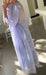 A person in the elegant Esperanza Maxi Dress — Classic by Noémiah, featuring puffed sleeves and a sheer lavender design, stands indoors near a sunlit area. They are holding a glass to their face with their back partially turned towards the camera, while shadows from a nearby window stretch across the floor.