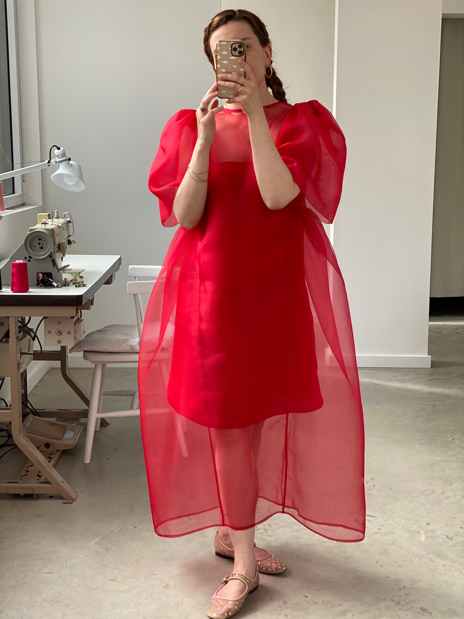 Wearing the Noémiah Esperanza Maxi Dress in a vivid sheer red with puffed sleeves, a person takes a selfie in a mirror. The room includes a sewing table equipped with a sewing machine and chair. They hold a phone adorned with a patterned case, standing gracefully on the concrete floor.