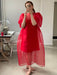 Wearing the Noémiah Esperanza Maxi Dress in a vivid sheer red with puffed sleeves, a person takes a selfie in a mirror. The room includes a sewing table equipped with a sewing machine and chair. They hold a phone adorned with a patterned case, standing gracefully on the concrete floor.