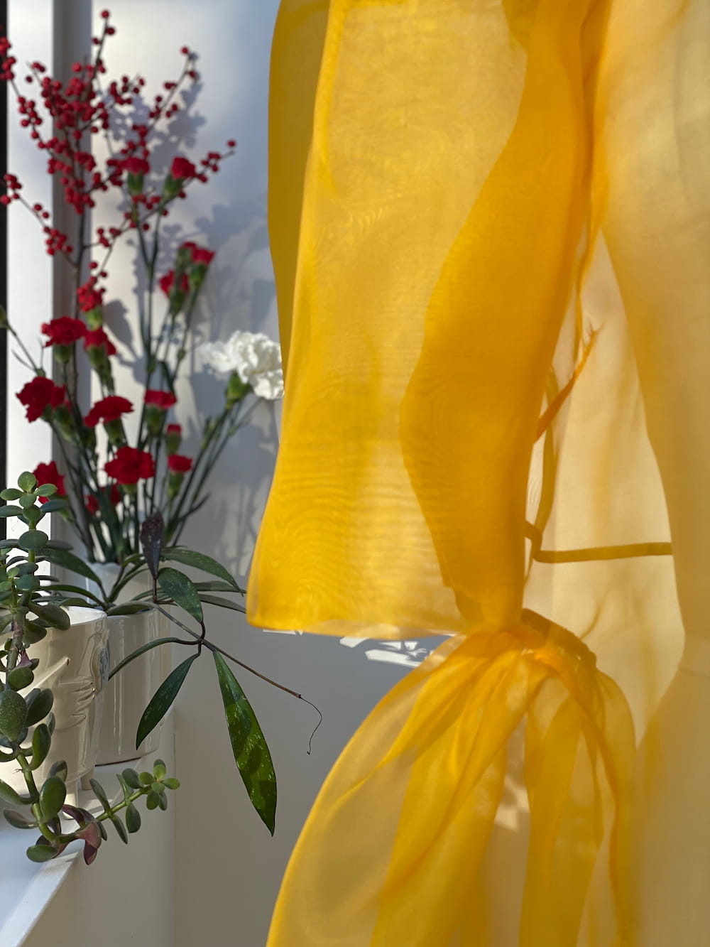 The Esperanza Maxi Dress by Noémiah, with its sheer yellow silk organza fabric, gently flutters in the sunlight alongside a vase filled with a vibrant bouquet of red and white flowers. The scene is harmoniously complemented by lush green leaves and soft lighting, enhancing its tranquility.