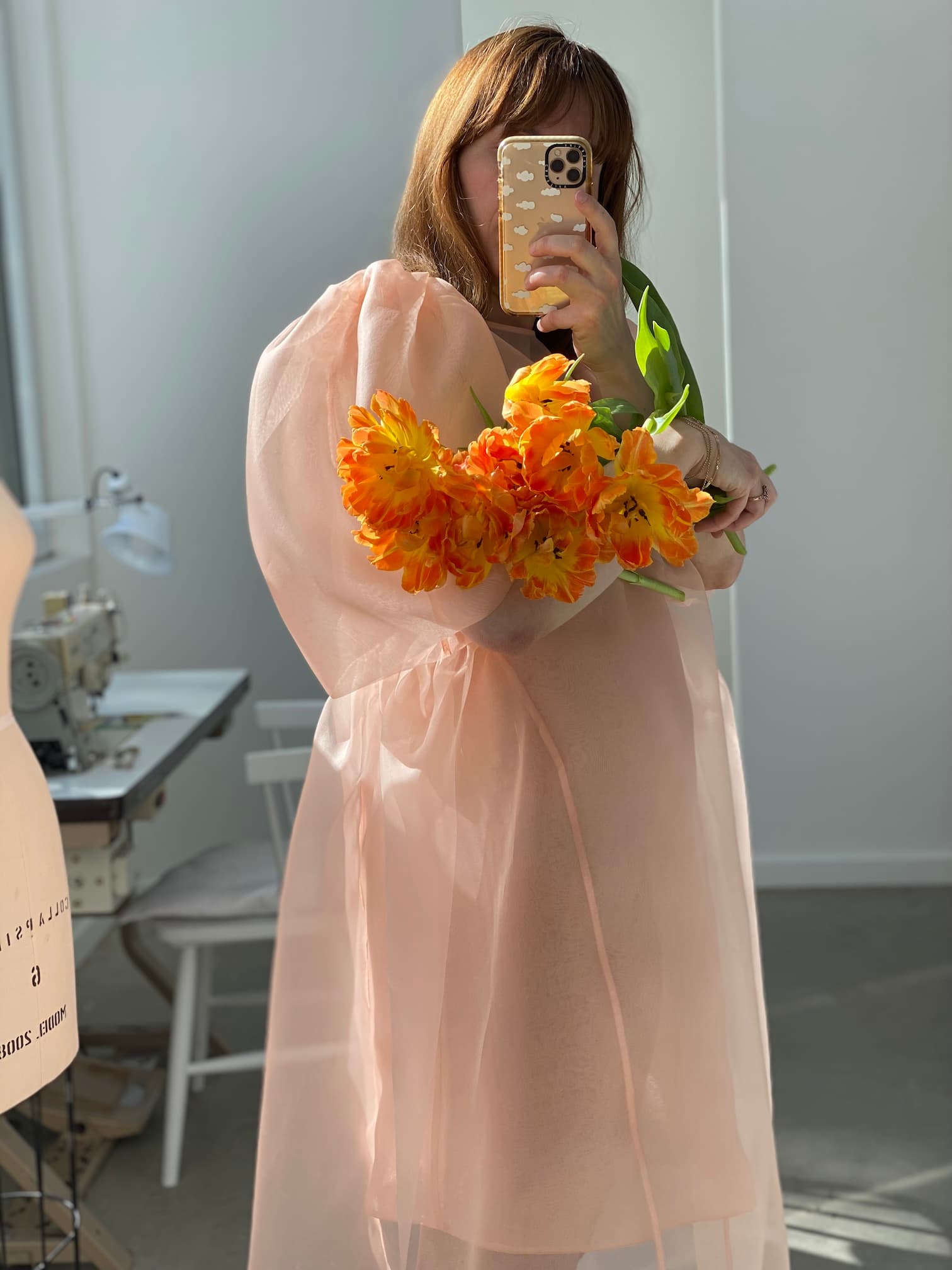 A person takes a mirror selfie holding a bouquet of bright orange flowers, dressed in the Noémiah Esperanza Maxi Dress — Classic, which features sheer fabric and elegant cutouts. In the background, a sewing machine and chair suggest a creative sewing or crafting space.