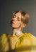 A woman with closed eyes, wearing the Esperanza Midi Dress in organza silk by Noémiah, stands against a gray background. The dress features sheer yellow fabric and puffed sleeves. She accessorizes with a white rose choker. Soft, natural light highlights her serene expression and subtle makeup, while her hair is styled in a loose updo.