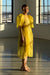 A person stands in a sunlit room wearing the elegant Esperanza Midi Dress by Noémiah, crafted from organza silk and featuring charming puffed sleeves. They are barefoot, with the sunlight casting shadows on the floor, while large windows frame an urban skyline outside.