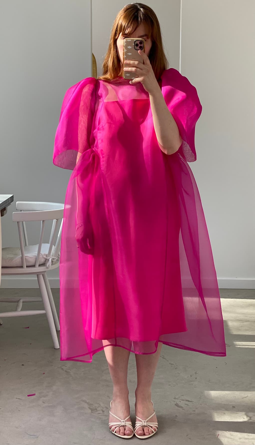 A person stands indoors taking a mirror selfie in the Noémiah Esperanza Midi Dress—an organza silk, bright pink dress with puffed sleeves and cutout detail—paired with white sandals. Their face is partially hidden by their phone; a white chair is on the left.