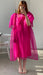 A person stands indoors taking a mirror selfie in the Noémiah Esperanza Midi Dress—an organza silk, bright pink dress with puffed sleeves and cutout detail—paired with white sandals. Their face is partially hidden by their phone; a white chair is on the left.