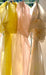 The Esperanza Midi Dress — Organza Silk by Noémiah, in shades of yellow, pale pink, and white, is shown hanging side by side in sunlight. The white dress features subtle yellow and orange stains, with a soft-focus window and greenery in the background.