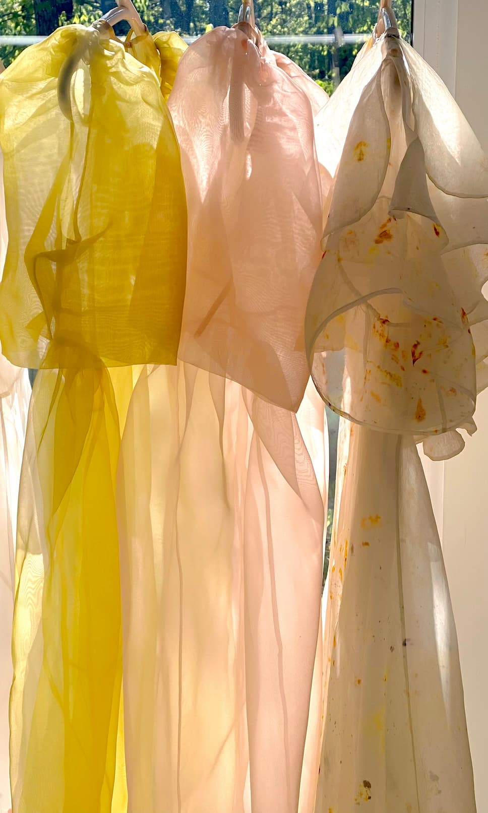 The Esperanza Midi Dress — Organza Silk by Noémiah, in shades of yellow, pale pink, and white, is shown hanging side by side in sunlight. The white dress features subtle yellow and orange stains, with a soft-focus window and greenery in the background.
