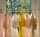Sheer pastel scarves in pink, white, yellow, and peach hang before a sunlit window, evoking the airy elegance of Noémiahs Esperanza Midi Dress in organza silk as green trees and sunlight stream through the glass.