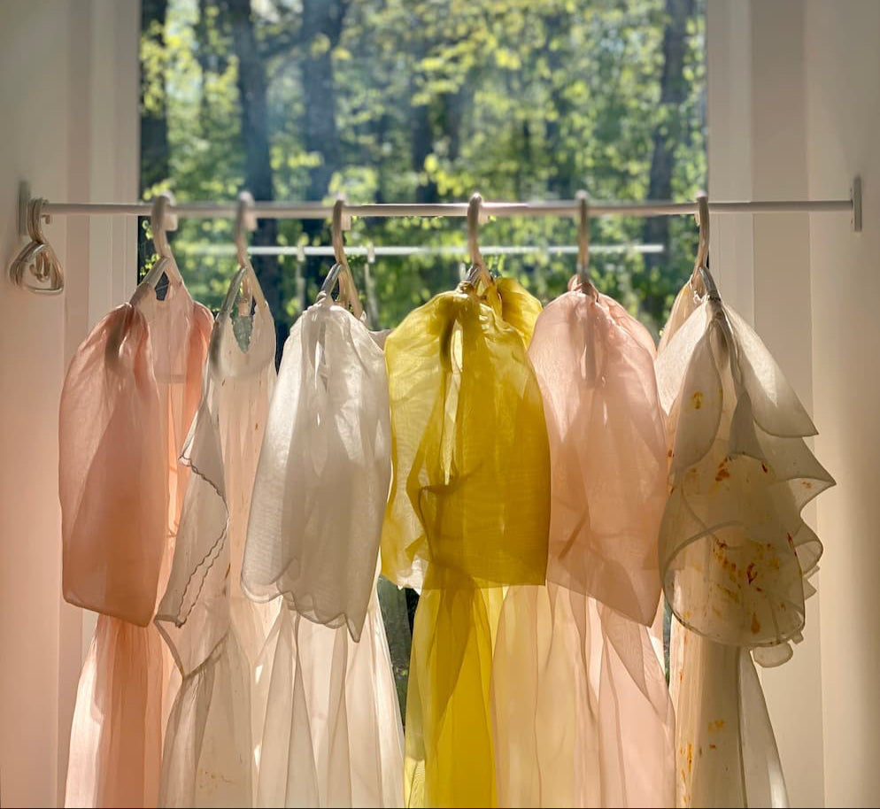 Sheer pastel scarves in pink, white, yellow, and peach hang before a sunlit window, evoking the airy elegance of Noémiahs Esperanza Midi Dress in organza silk as green trees and sunlight stream through the glass.