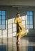 An individual wearing the Noémiah Esperanza Midi Dress, made from organza silk, stands barefoot in a sunlit room with large windows. The flowing yellow dress features puffed sleeves and casts long shadows on the polished floor as it catches the light, creating a delicate, airy effect.