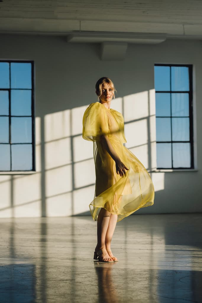 An individual wearing the Noémiah Esperanza Midi Dress, made from organza silk, stands barefoot in a sunlit room with large windows. The flowing yellow dress features puffed sleeves and casts long shadows on the polished floor as it catches the light, creating a delicate, airy effect.