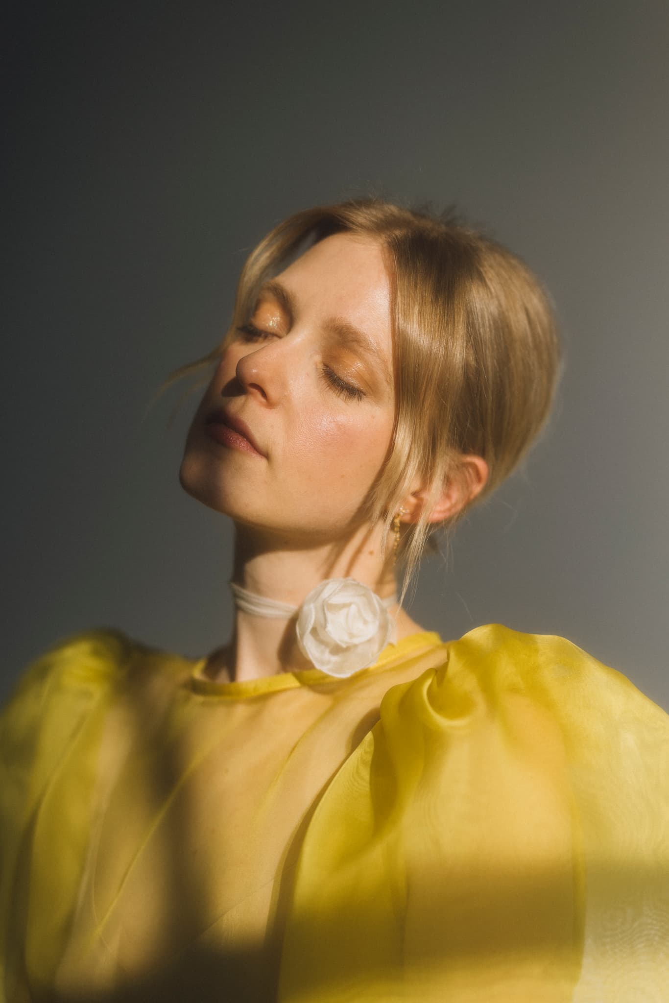 A woman with closed eyes, wearing the Esperanza Midi Dress in organza silk by Noémiah, stands against a gray background. The dress features sheer yellow fabric and puffed sleeves. She accessorizes with a white rose choker. Soft, natural light highlights her serene expression and subtle makeup, while her hair is styled in a loose updo.