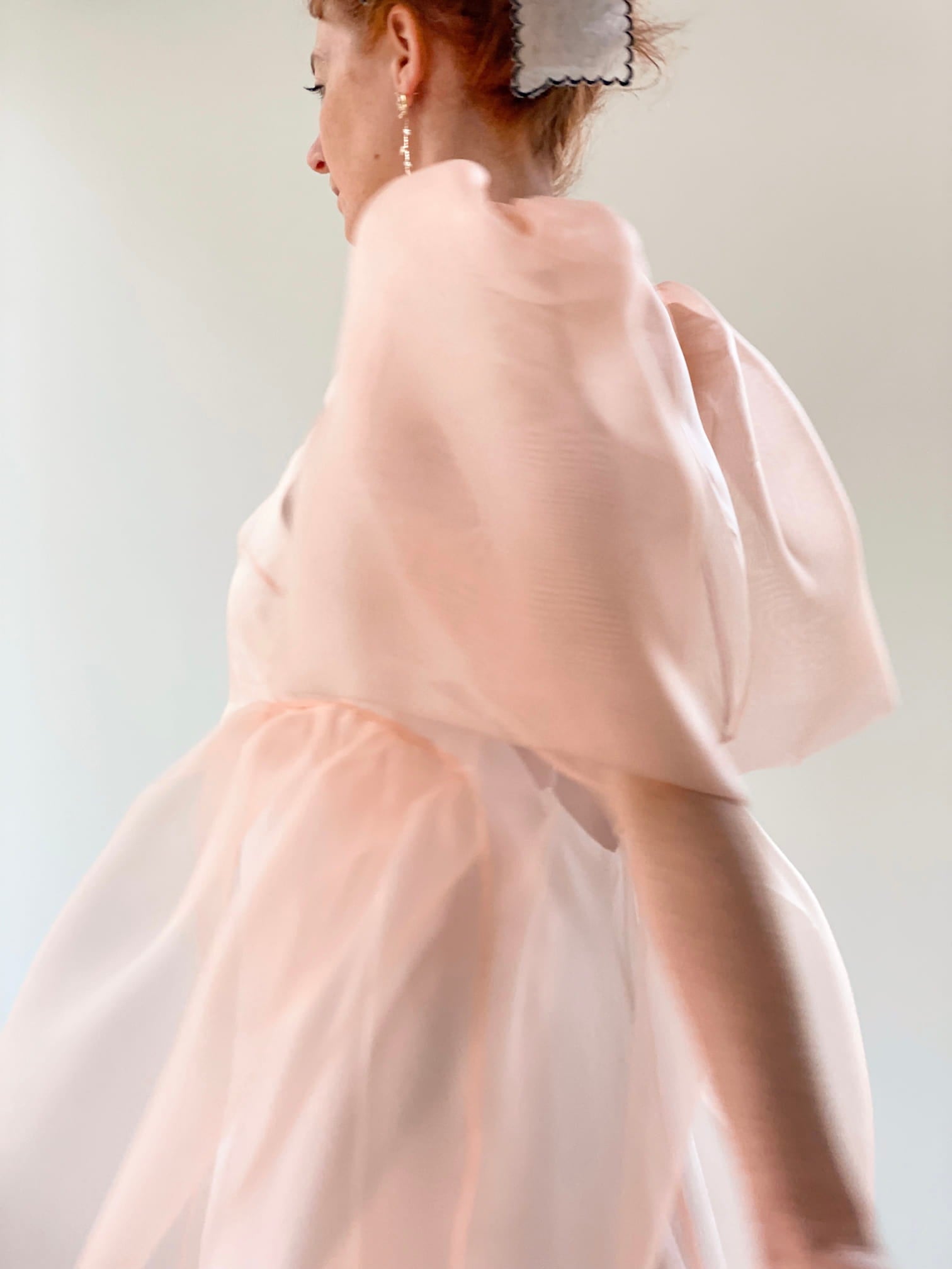 The image depicts an individual in motion clad in the Esperanza Midi Dress — Organza Silk by Noémiah, a flowy attire in soft pink with puffed sleeves and a delicate hair accessory. The minimalistic, neutral background accentuates the elegance of the dress while capturing the graceful movement of the fabric.