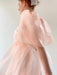 The image depicts an individual in motion clad in the Esperanza Midi Dress — Organza Silk by Noémiah, a flowy attire in soft pink with puffed sleeves and a delicate hair accessory. The minimalistic, neutral background accentuates the elegance of the dress while capturing the graceful movement of the fabric.