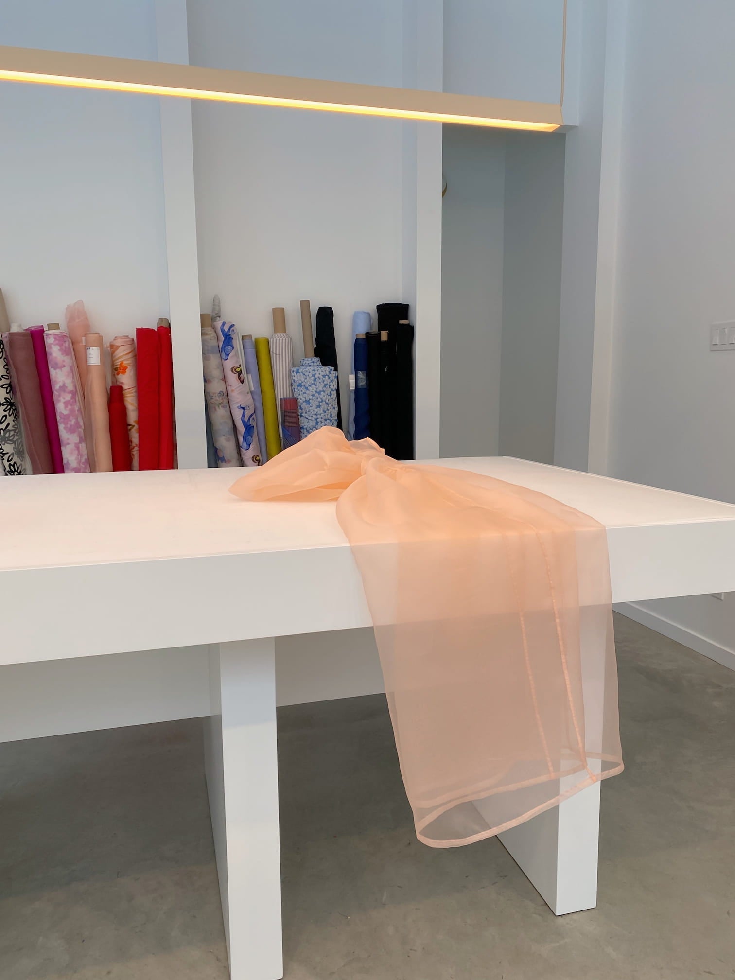 A peach-colored sheer fabric, reminiscent of the Esperanza Midi Dress made from organza silk by Noémiah, is draped over a white table in a bright room. Rolls of various colorful fabrics stand vertically against the wall in the background, suggesting future creations such as dresses with puffed sleeves.