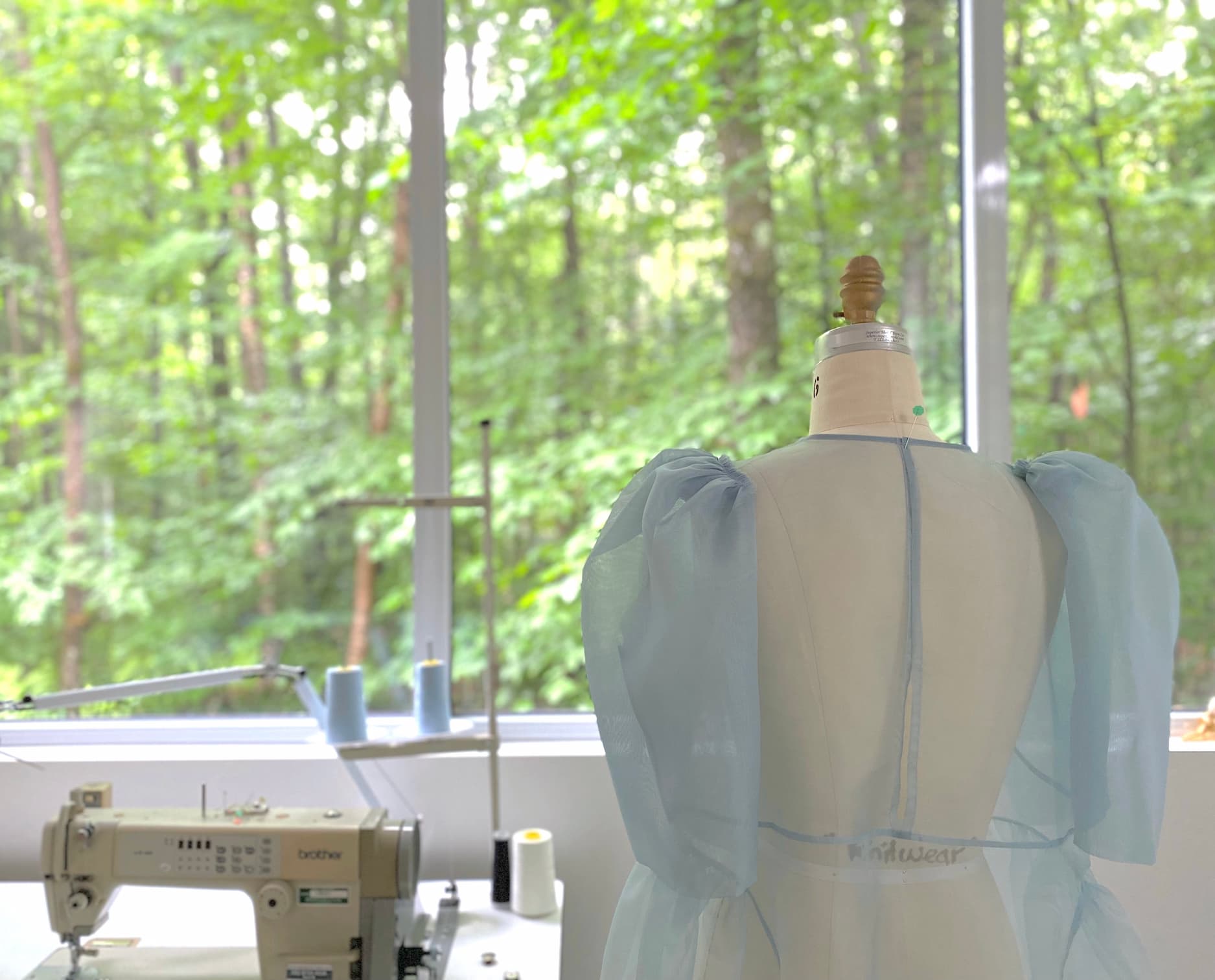 An Esperanza Midi Dress — Organza Silk by Noémiah, showcasing a light blue and sheer design with puffed sleeves, is displayed on a dress form facing large windows overlooking a lush green forest. A sewing machine and thread spools are arranged on the table, creating a serene and creative studio atmosphere.