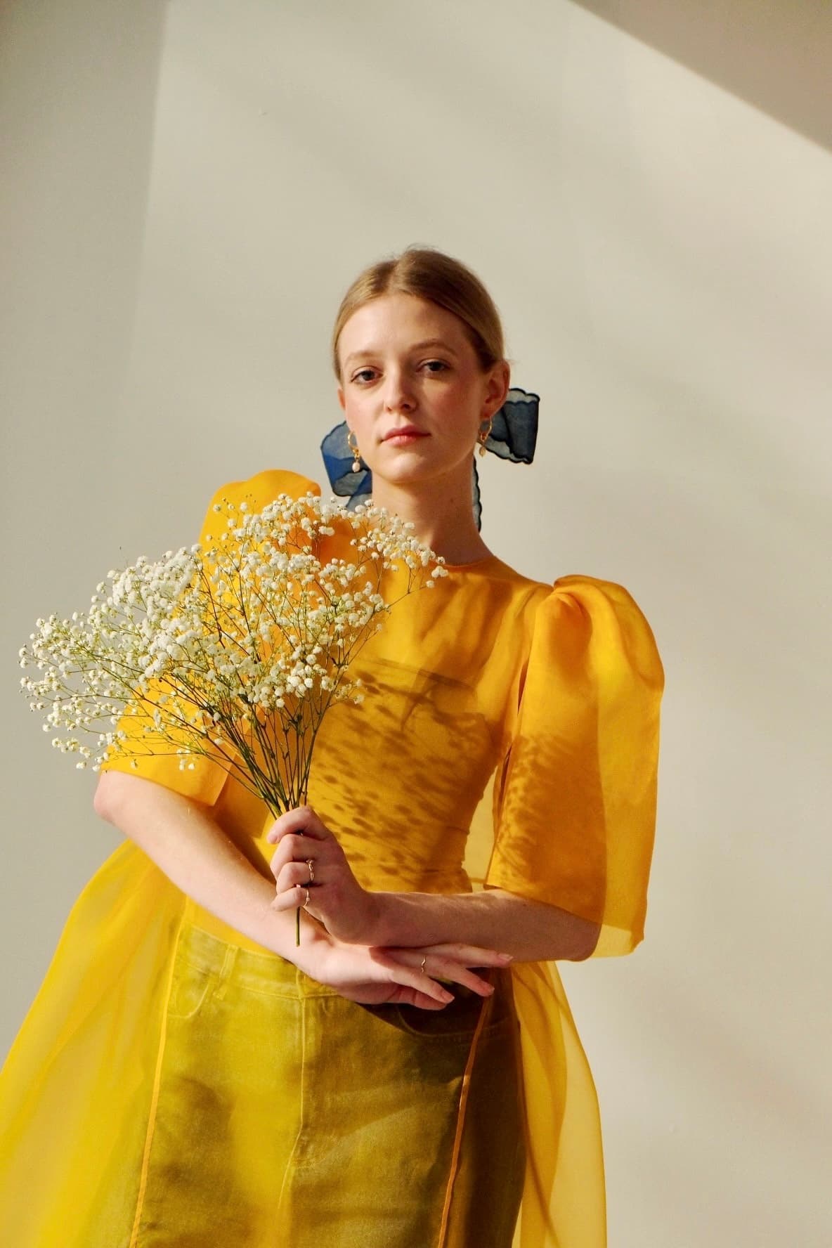 A woman wearing the Esperanza Midi Dress in yellow organza silk by Noémiah, featuring puffed sleeves, holds a bouquet of small white flowers. She has blue ribbons in her hair and stands against a softly shadowed light-colored background.